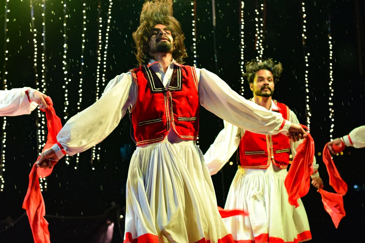 A performer in a white fustanella-style costume with a red sash, mid-turn on a lit stage — representative of Greek folk-dance costume, not this show's troupe