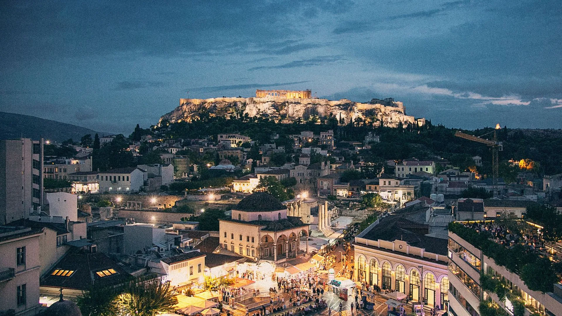 Athens' old town glowing at dusk below the floodlit Acropolis, with a lit rooftop taverna terrace in the foreground — representative of the city the evening happens in, not the taverna itself