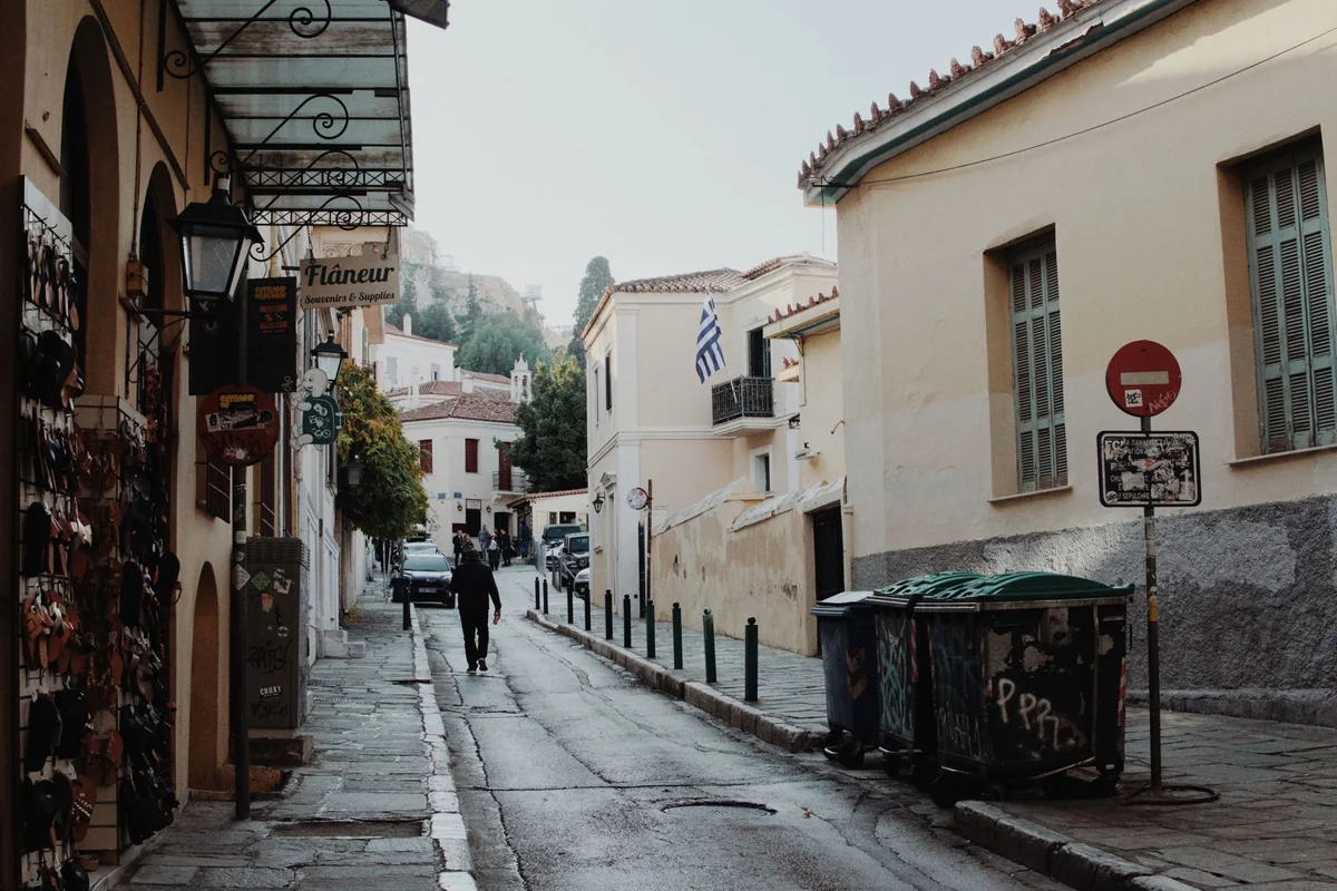 A narrow Plaka lane with a Greek flag and the Acropolis rock visible up the hill — representative of the district's lanes